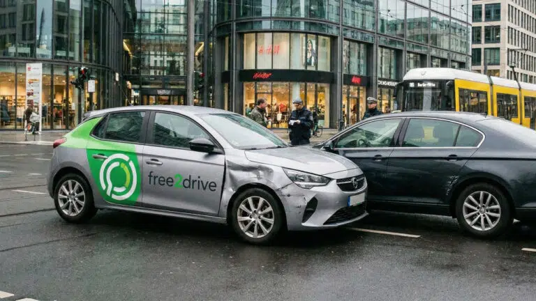 Beschädigtes Carsharing-Fahrzeug nach einer Kollision, im Hintergrund Polizei und Straßenbahn, Symbolbild für einen Unfall mit einem geliehenen Auto