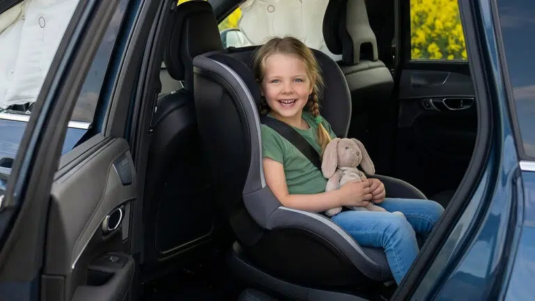 Lächelndes Mädchen sitzt angeschnallt mit Kuscheltier in einem Kindersitz auf der Rückbank, Symbolbild für den Austausch nach einem Unfall